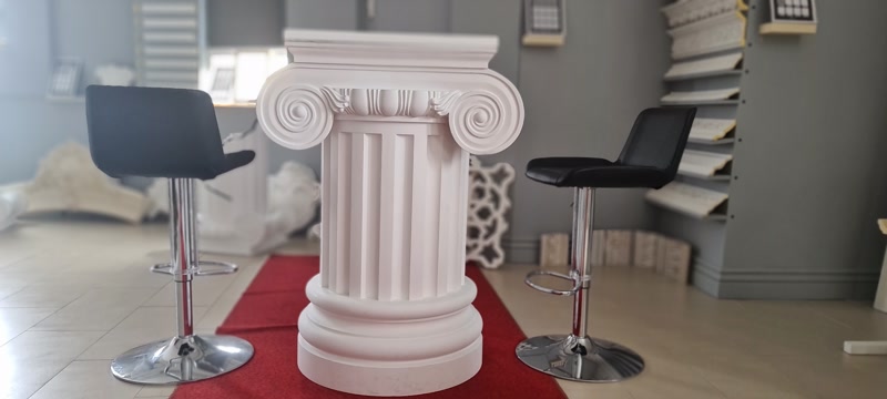 Kırmızı halı üzerinde sergilenen beyaz poliüretan iyonik sütun ve etrafındaki siyah krom ayaklı bar tabureleri.