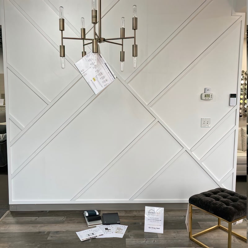 Çapraz ve geometrik çıtalama yapılmış beyaz duvar önünde pirinç avize ve siyah puf ile modern salon tasarımı.