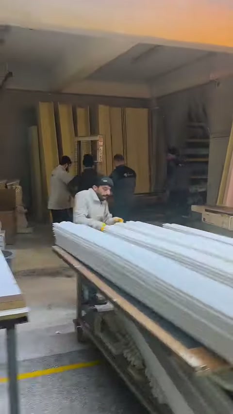 Çalışanlar, uzun beyaz profilleri geniş üretim masasının üzerinde düzenliyor.