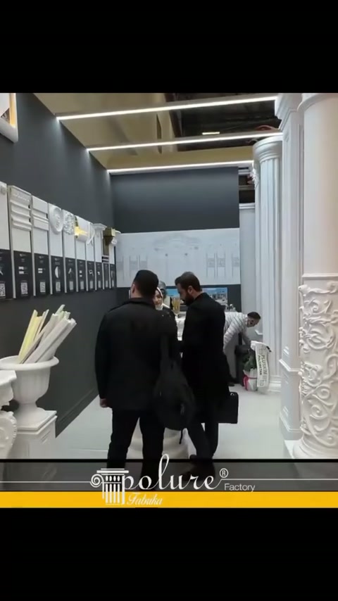 Stand koridorunda iki ziyaretçi, profillerle kaplı duvarı değerlendiriyor
